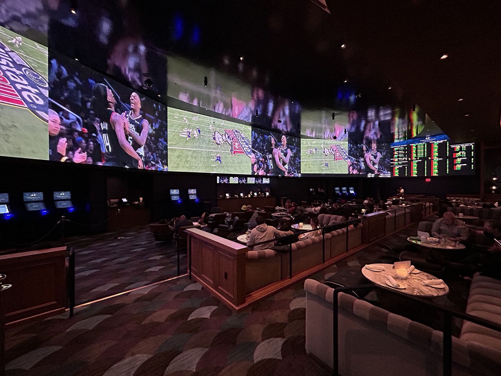 The sportsbook at Fontainebleau Las Vegas: a curved video wall showing football and basketball above rows of lounge seating, with the odds boards at right.